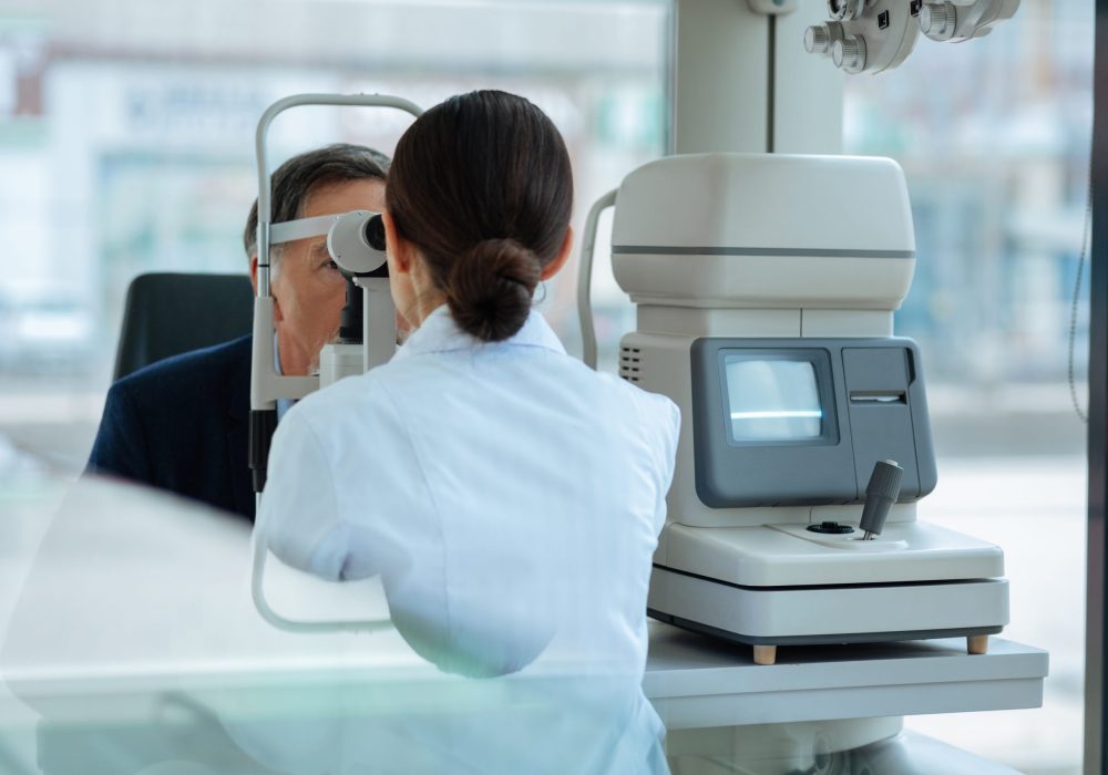 Nice dark haired woman using the optical instrument while checking the eyesight of her patient